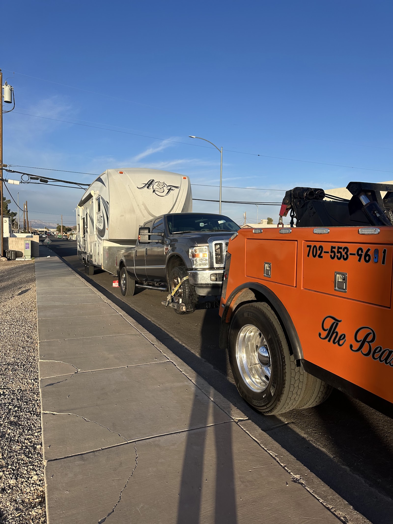 Orange wrecker towing a pickup pulling a fifth-wheel RV, with phone number 702-553-9691 visible