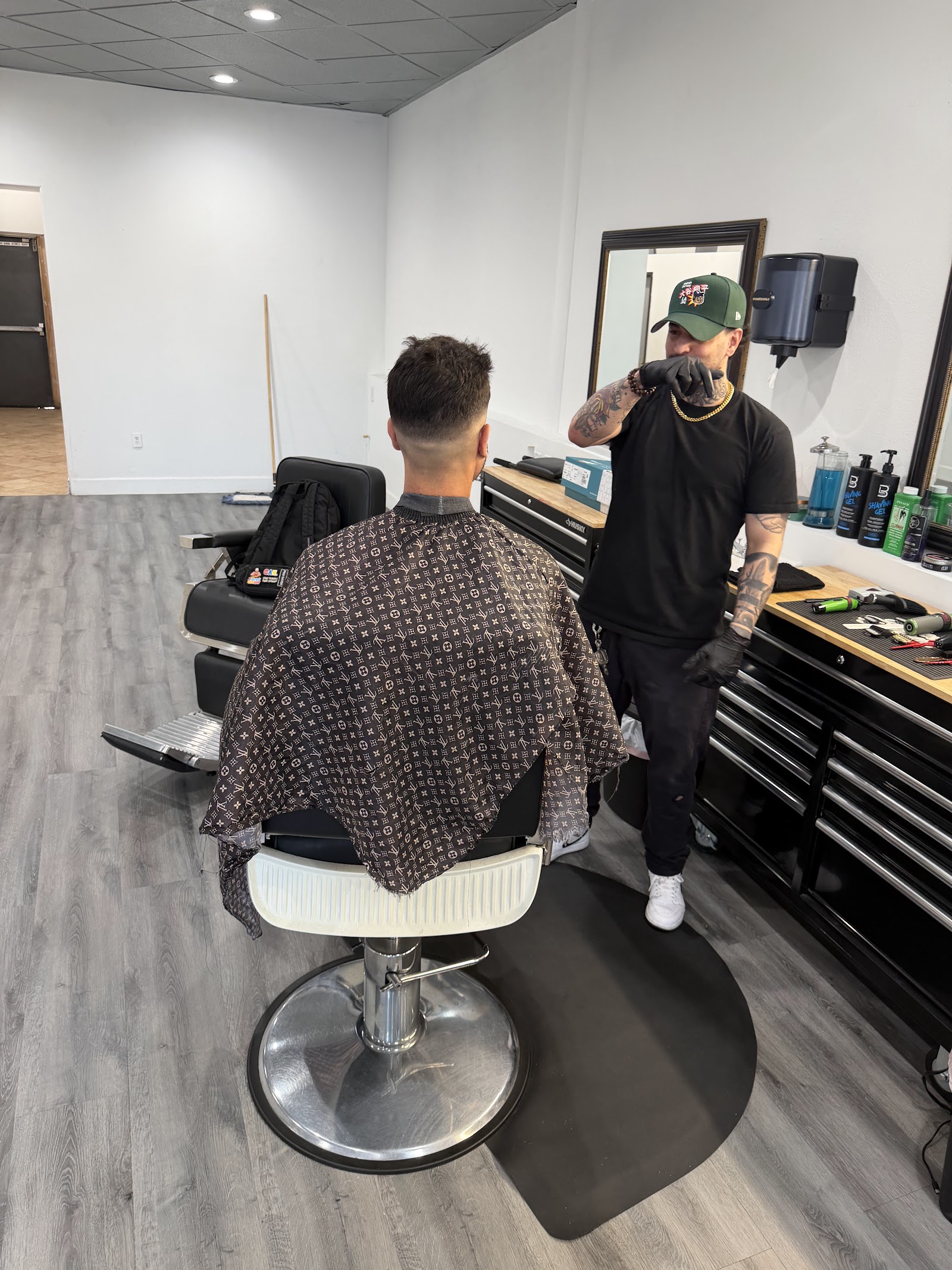 Barber in green cap standing near workstation while client sits in chair with patterned cape