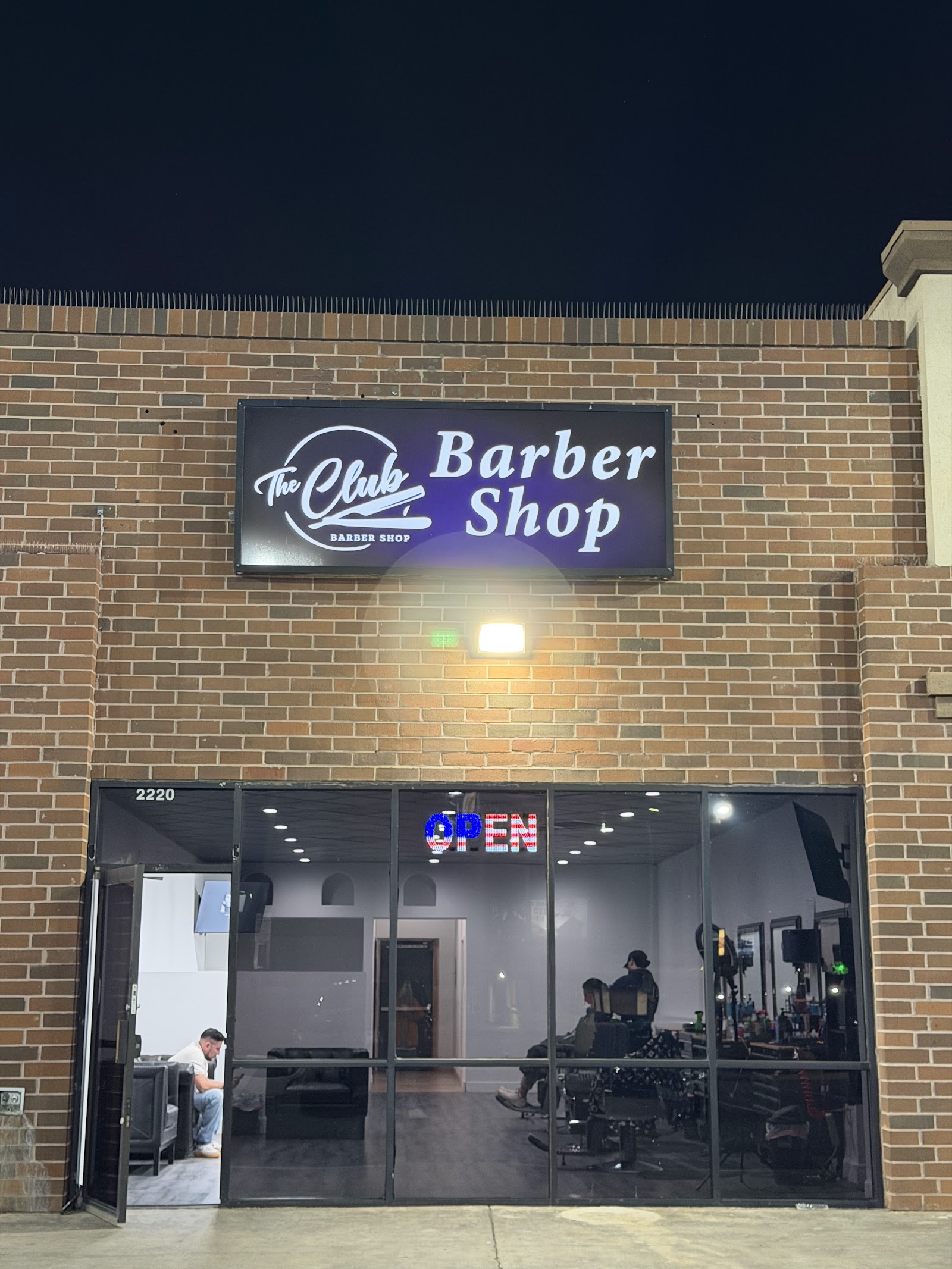 The Club Barber Shop illuminated storefront sign at night with OPEN sign and brick facade