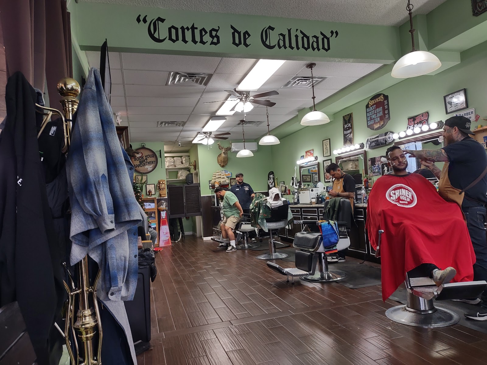 Busy barbershop interior with 'Cortes de Calidad' wall text, several barbers and clients