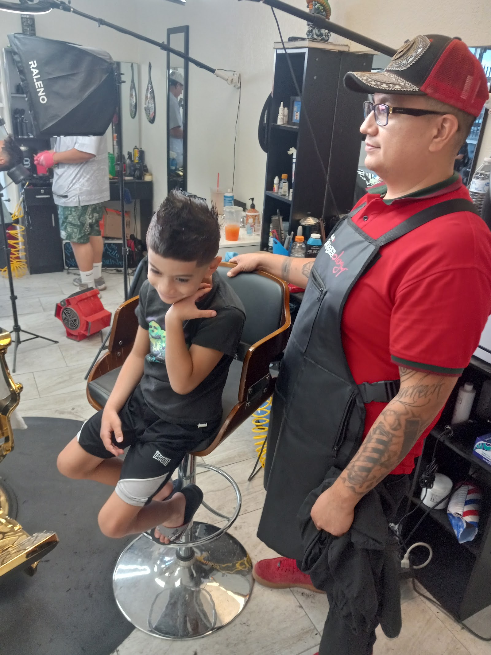 Barber in red shirt and apron standing beside young boy with spiky haircut in chair
