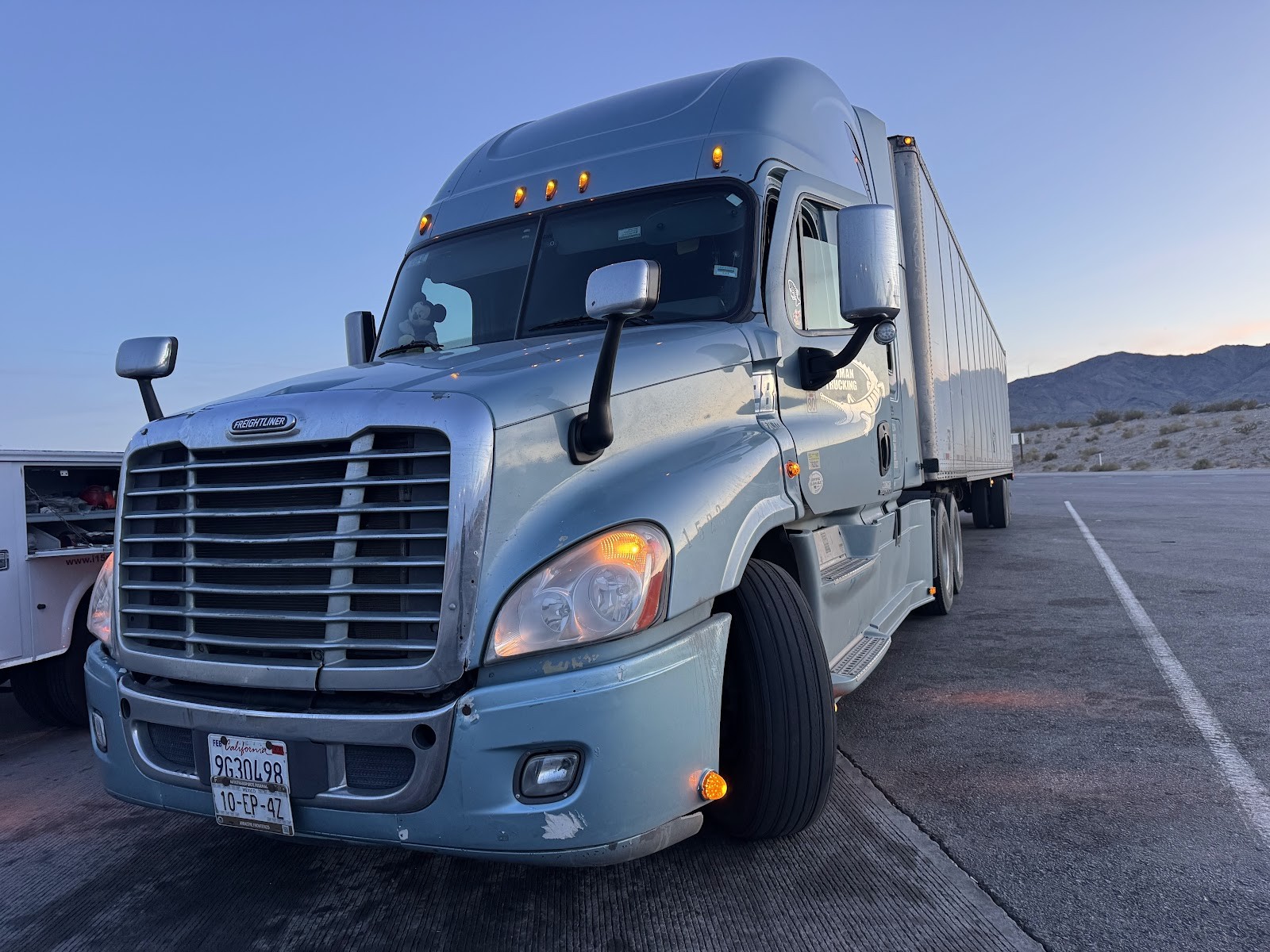 Silver Freightliner Cascadia semi-truck with trailer parked at dusk in the desert