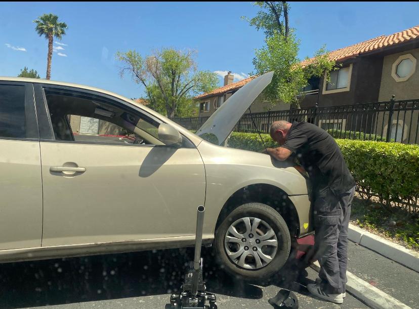 Mechanic working under the hood of a silver sedan on a jack in a residential parking lot