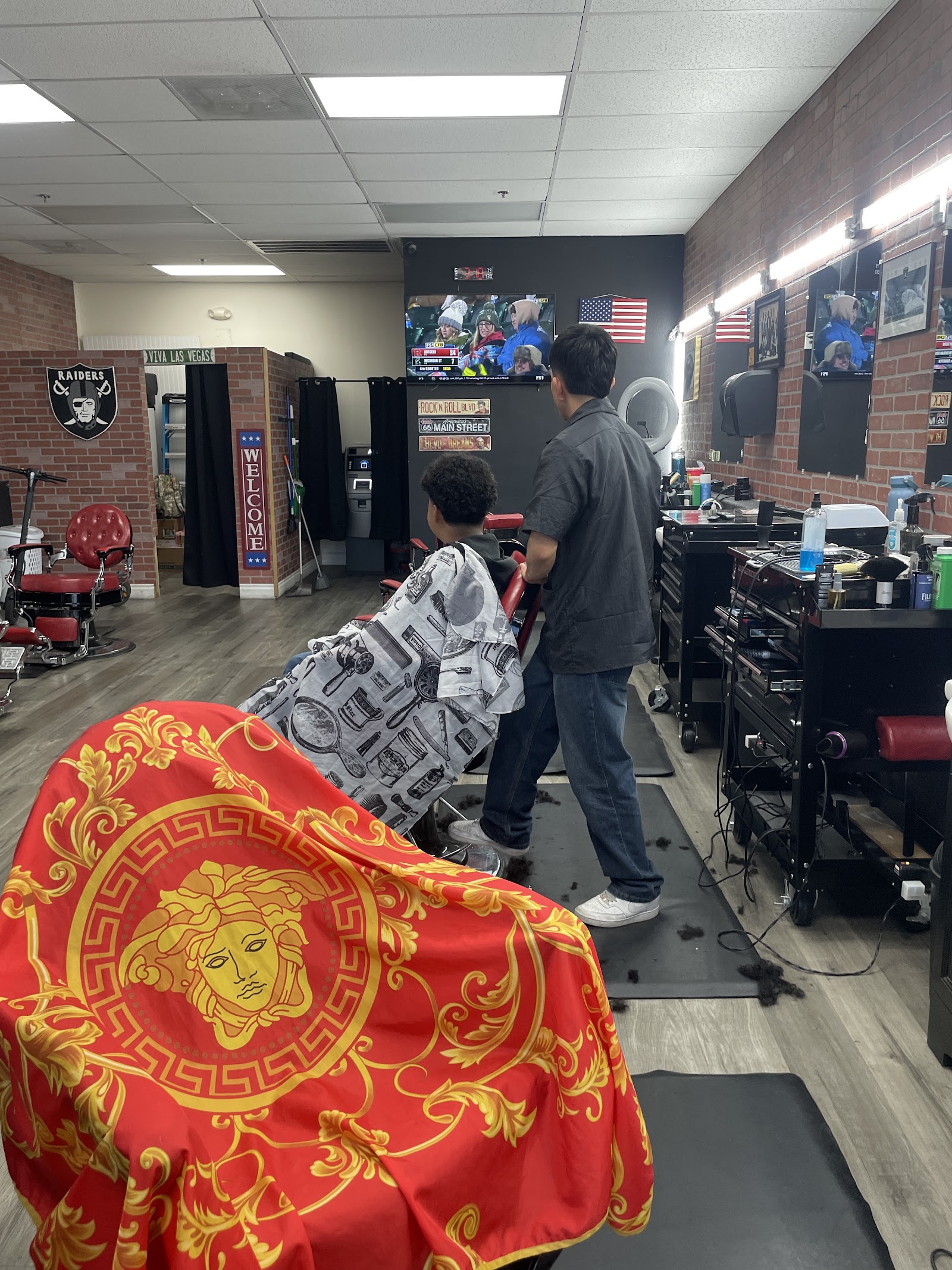 Barber cutting a young client's hair inside the shop with TVs and Raiders decor