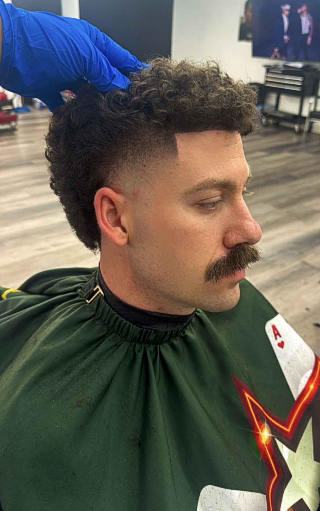 Man with curly mullet, taper fade, and mustache being styled by gloved barber