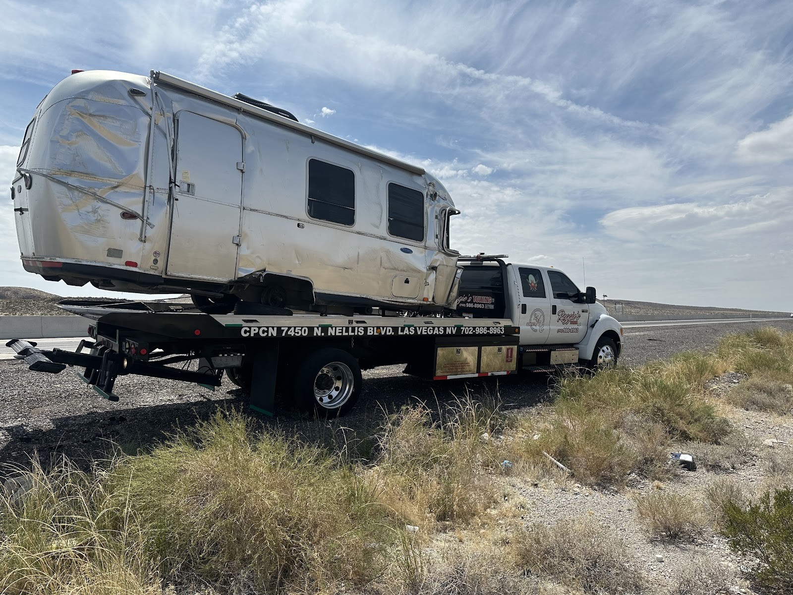 Reggie's Towing flatbed hauling a damaged Airstream trailer along a desert highway