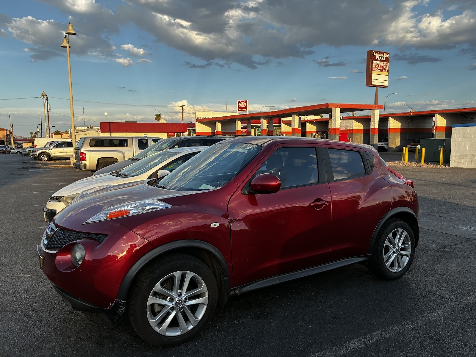 Red Nissan Juke parked in a lot at golden hour with plaza and gas station in background