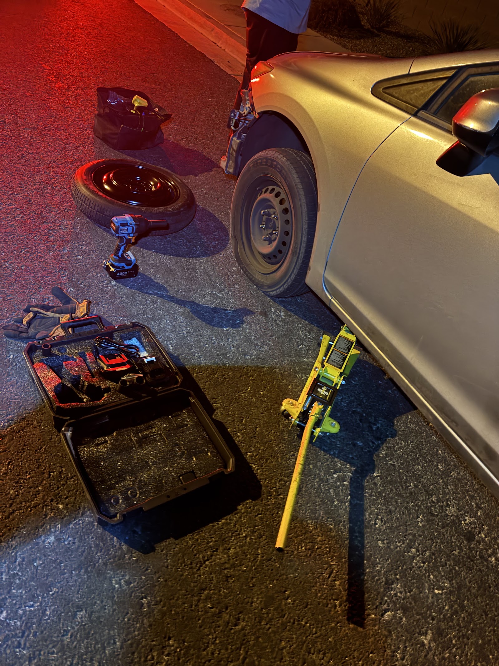 Nighttime tire change with spare, impact wrench and floor jack beside a silver sedan