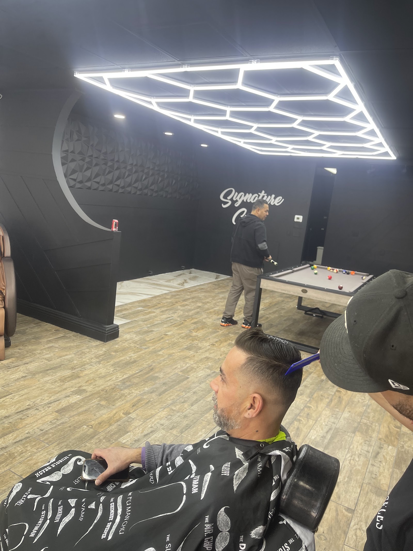 Barbershop interior with hexagonal LED lighting, pool table and Signature Cuts wall sign, client getting a haircut