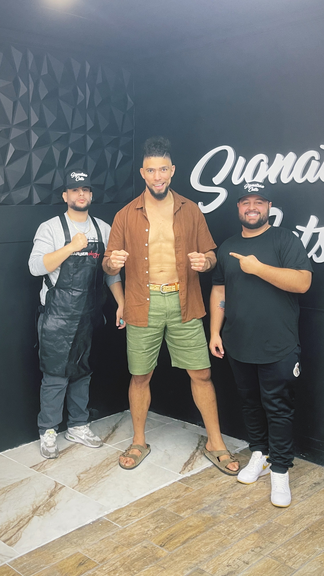 Two barbers posing with a client in front of the Signature Cuts wall sign